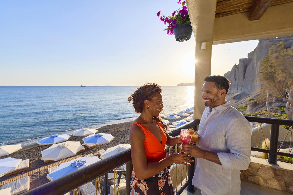 Couple enjoying drinks at Royal Beach Club Santorini with black sand beach and Aegean Sea views