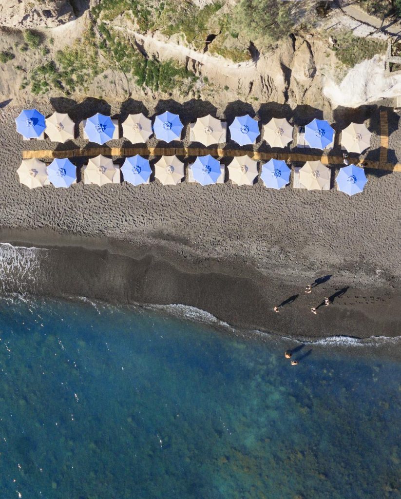 Aerial view of Royal Beach Club Santorini black sand beach blue umbrellas and wooden walkways