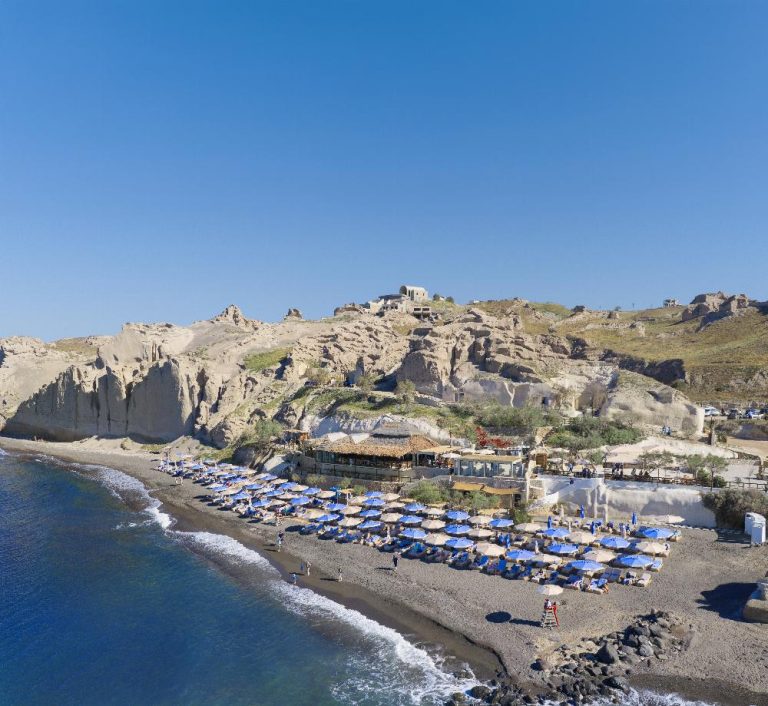 Aerial view of Royal Beach Club Santorini black sand beach with volcanic cliffs and Aegean Sea