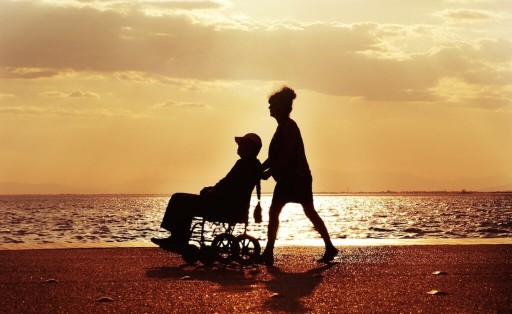Silhouette of a person pushing a wheelchair user along the shore at sunset near a cruise port