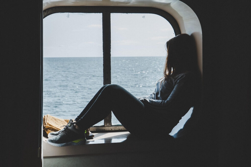 Woman sitting alone by a ship window looking out at the ocean — cruising with anxiety and panic disorder